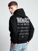 Black graphic hoodie worn by model with white Monaco branding and oversized lifestyle text on back.